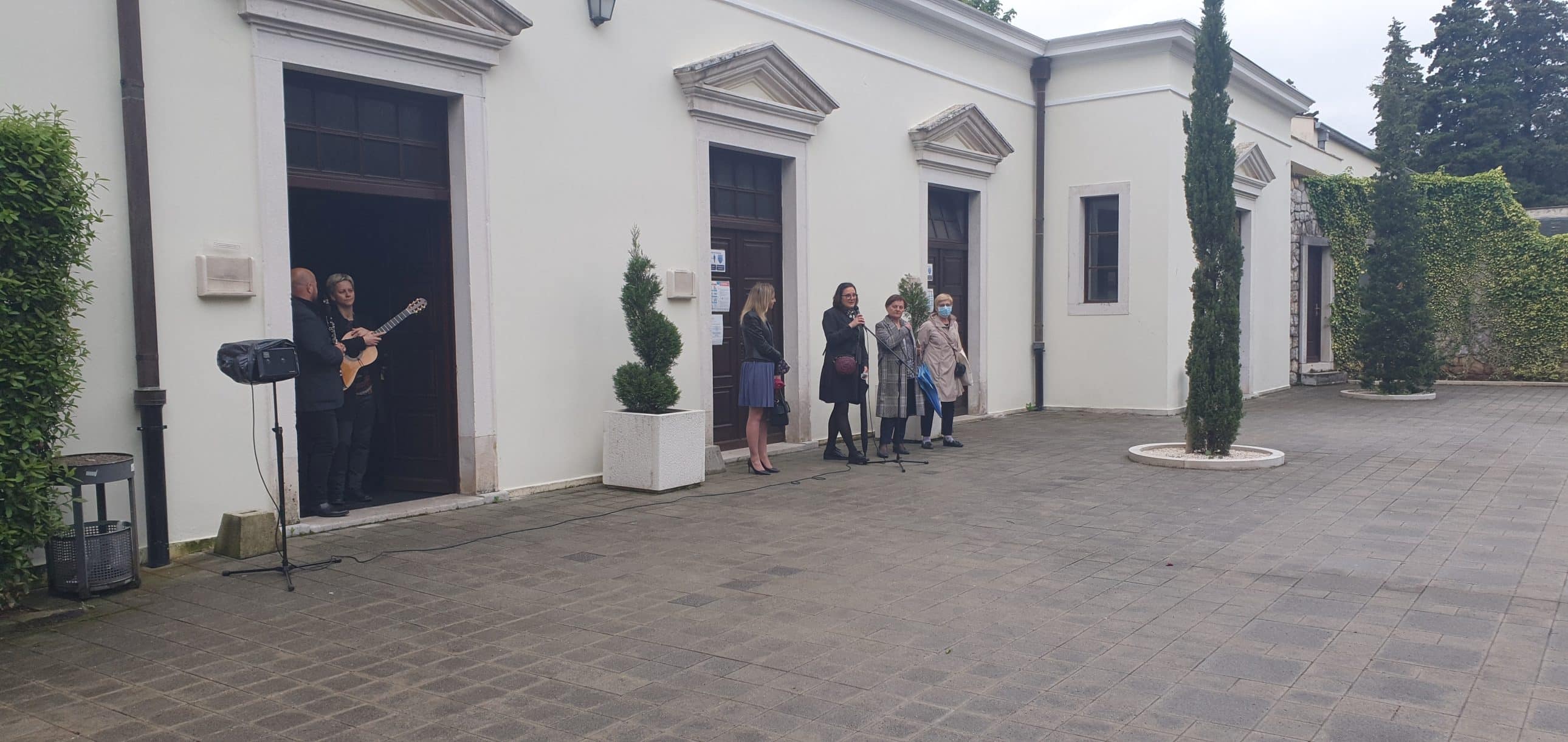 Glazbenik s gitarom i okupljeni građani ispred oproštajne dvorane na groblju.