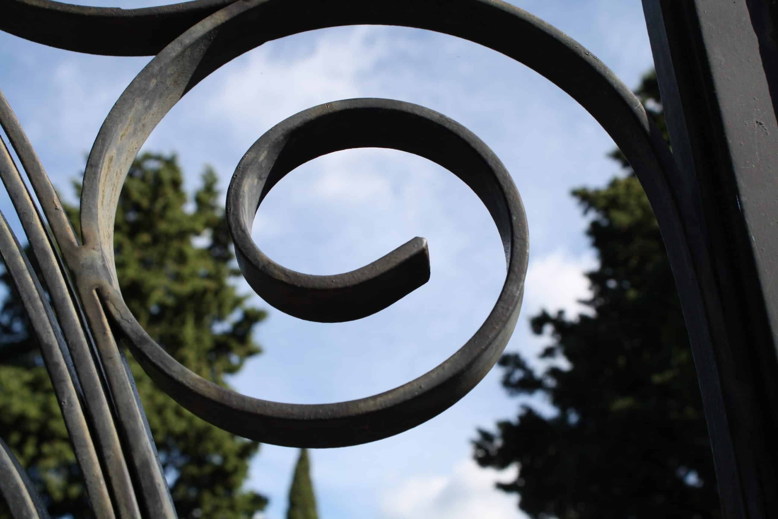 Kovani spiralni ukras željezne ograde na groblju kroz koji se vide čempresi i plavo nebo.