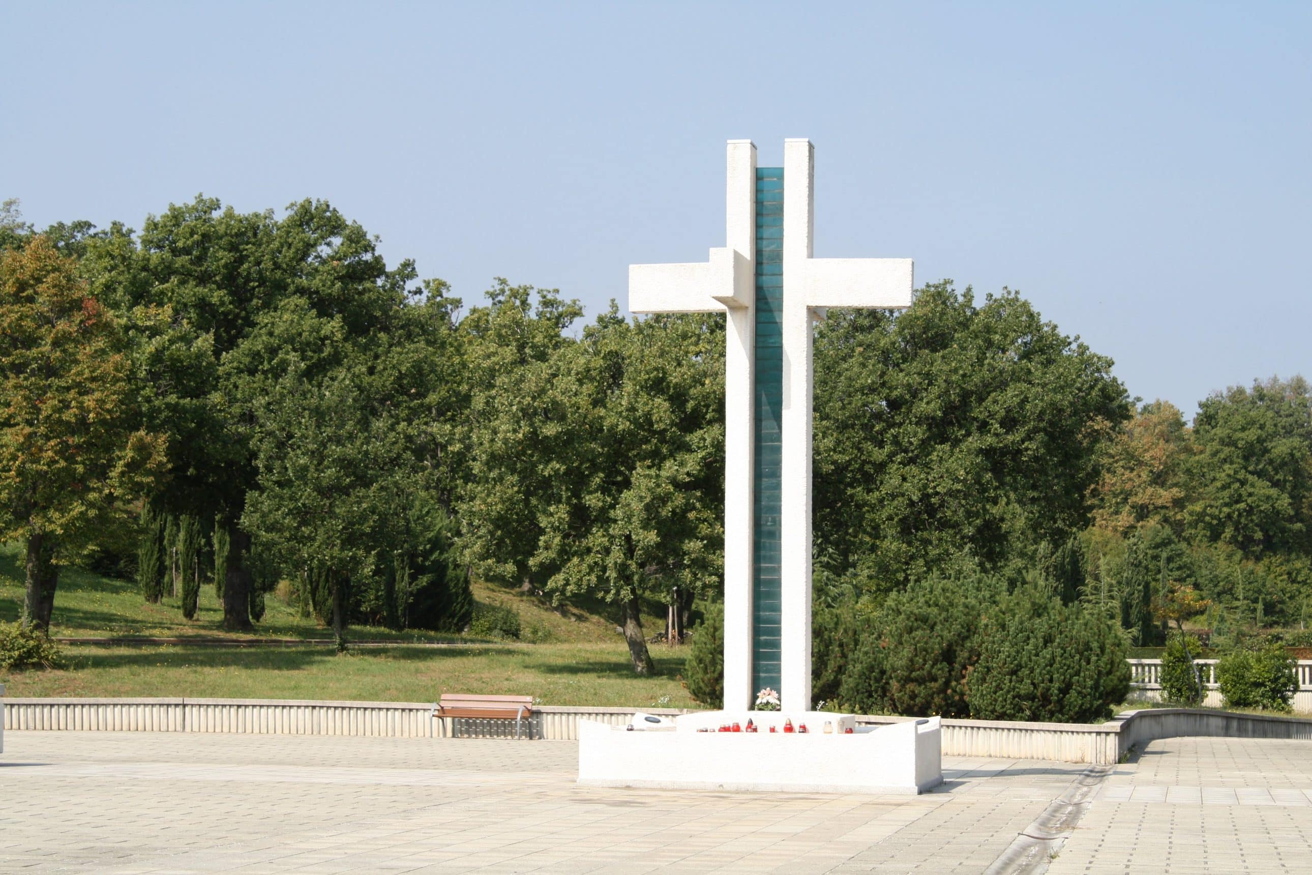 Veliki bijeli spomen-križ sa staklenim detaljem na popločanom platou groblja okružen zelenilom.