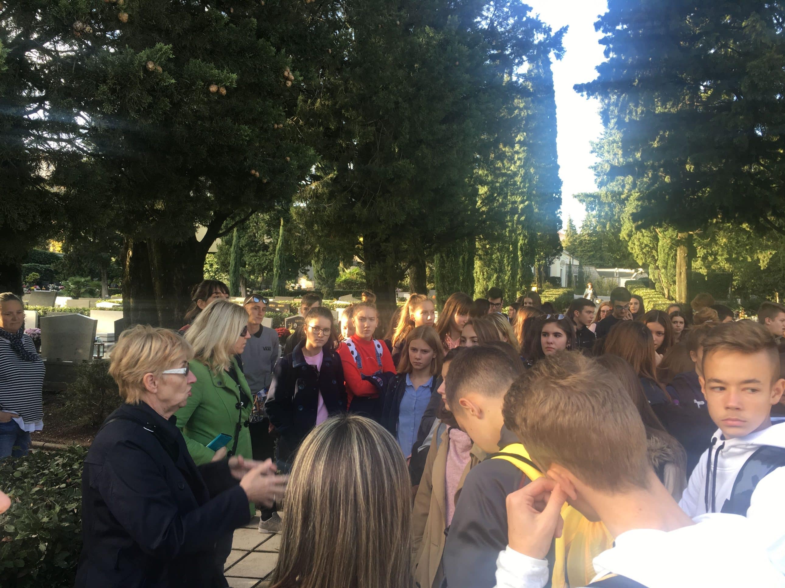 Skupina učenika sluša vodiča tijekom edukativnog obilaska na groblju među visokim čempresima.