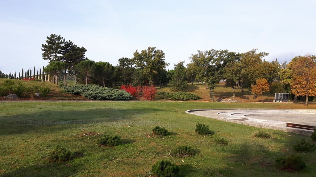 Uređeni park i zelene površine na Centralnom gradskom groblju Drenova s drvećem i popločanim trgom.