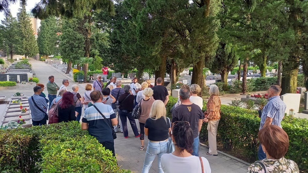Grupa posjetitelja na vođenom obilasku stazom povijesnog groblja okruženog zelenilom i spomenicima.