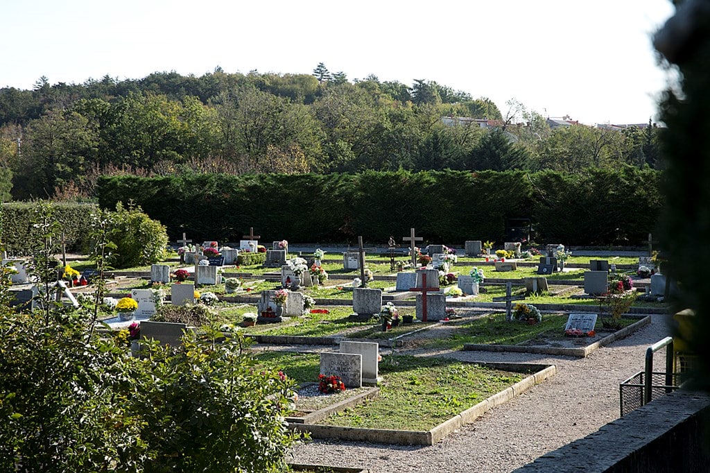 Uređeno groblje s nadgrobnim spomenicima, križevima i cvijećem okruženo zelenilom i drvećem.